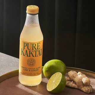 A glass bottle of Pure Kaktai Lime & Ginger Sparkling Cactus Water on a tray next to a lime and ginger root.
